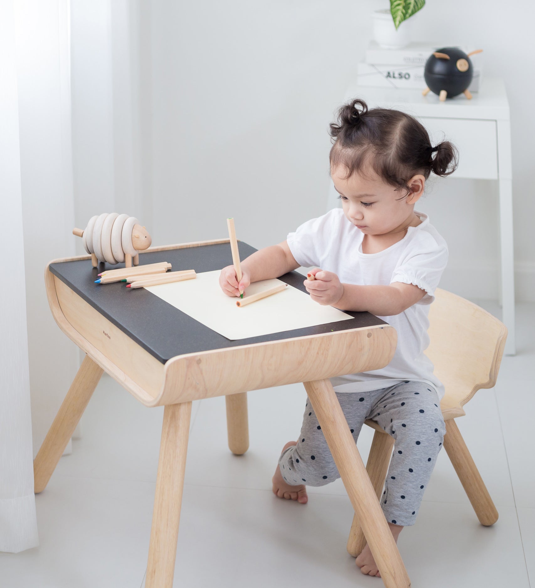 A child sitting by a PlanToys Black Table & Chair. The child is writing and a piece of paper. 