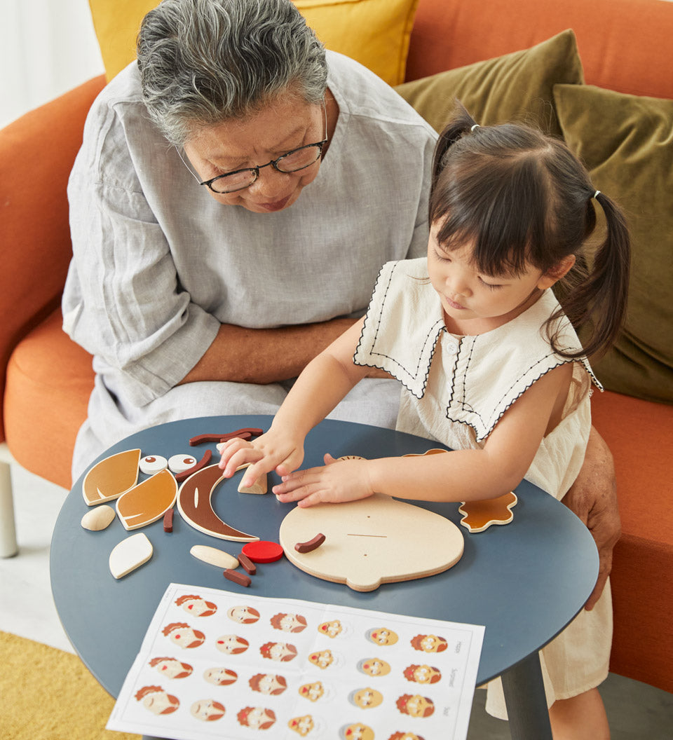 Young girl and woman sat at a round table playing with the PlanToys wooden build a face toy