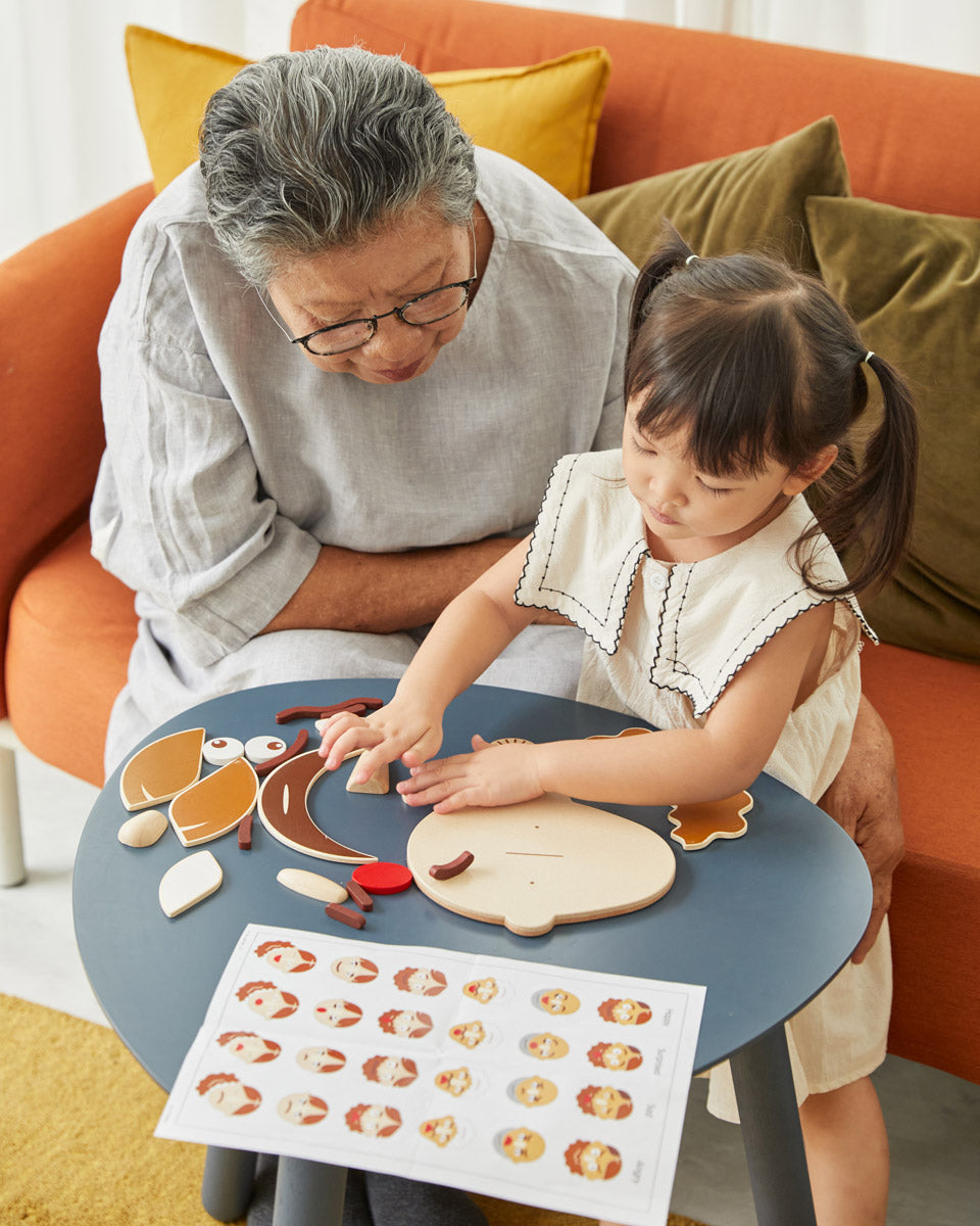 Young girl and woman sat at a round table playing with the PlanToys wooden build a face toy