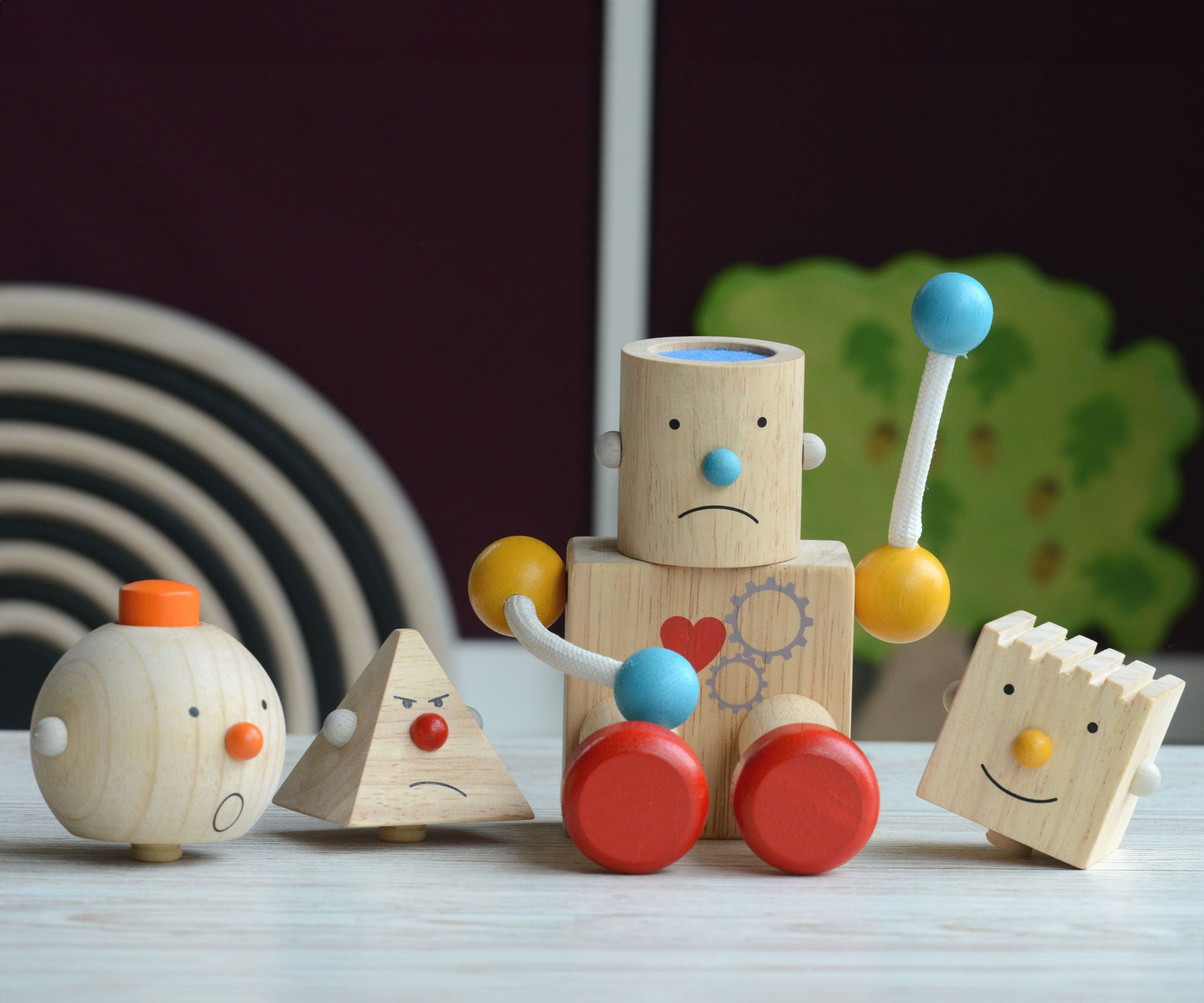 The PlanToys Build a Robot placed on a wooden surface n front of other wooden toys. The robot has it's cylinder shaped head on with a sad expression. 