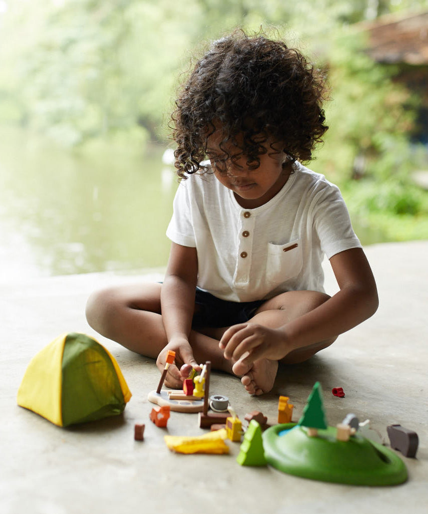 A child sitting on a floor playing with the PlanToys PlanWorld Camping Set.
