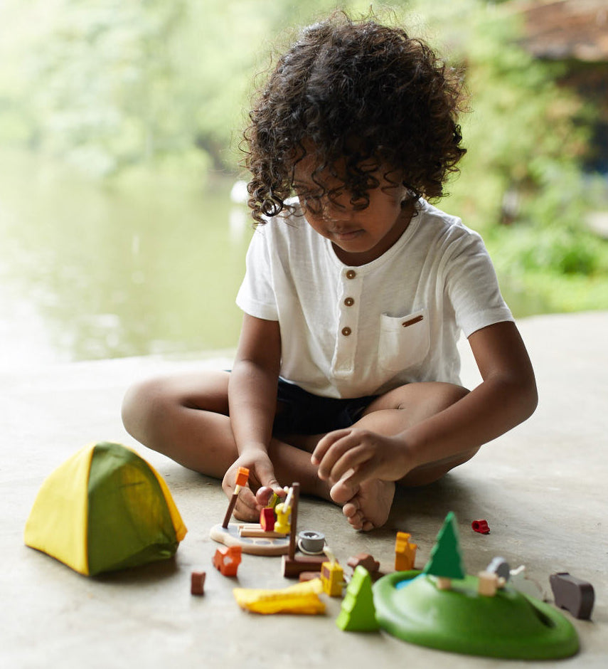 A child sitting on a floor playing with the PlanToys PlanWorld Camping Set.
