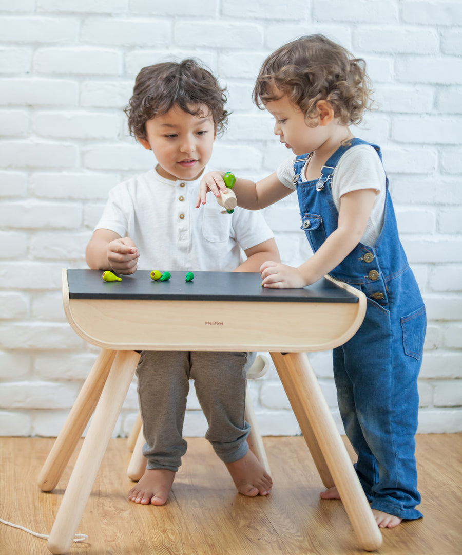 A child sitting by a PlanToys Black Table & Chair. Another child is standing by their side.