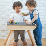 A child sitting by a PlanToys Black Table & Chair. Another child is standing by their side.