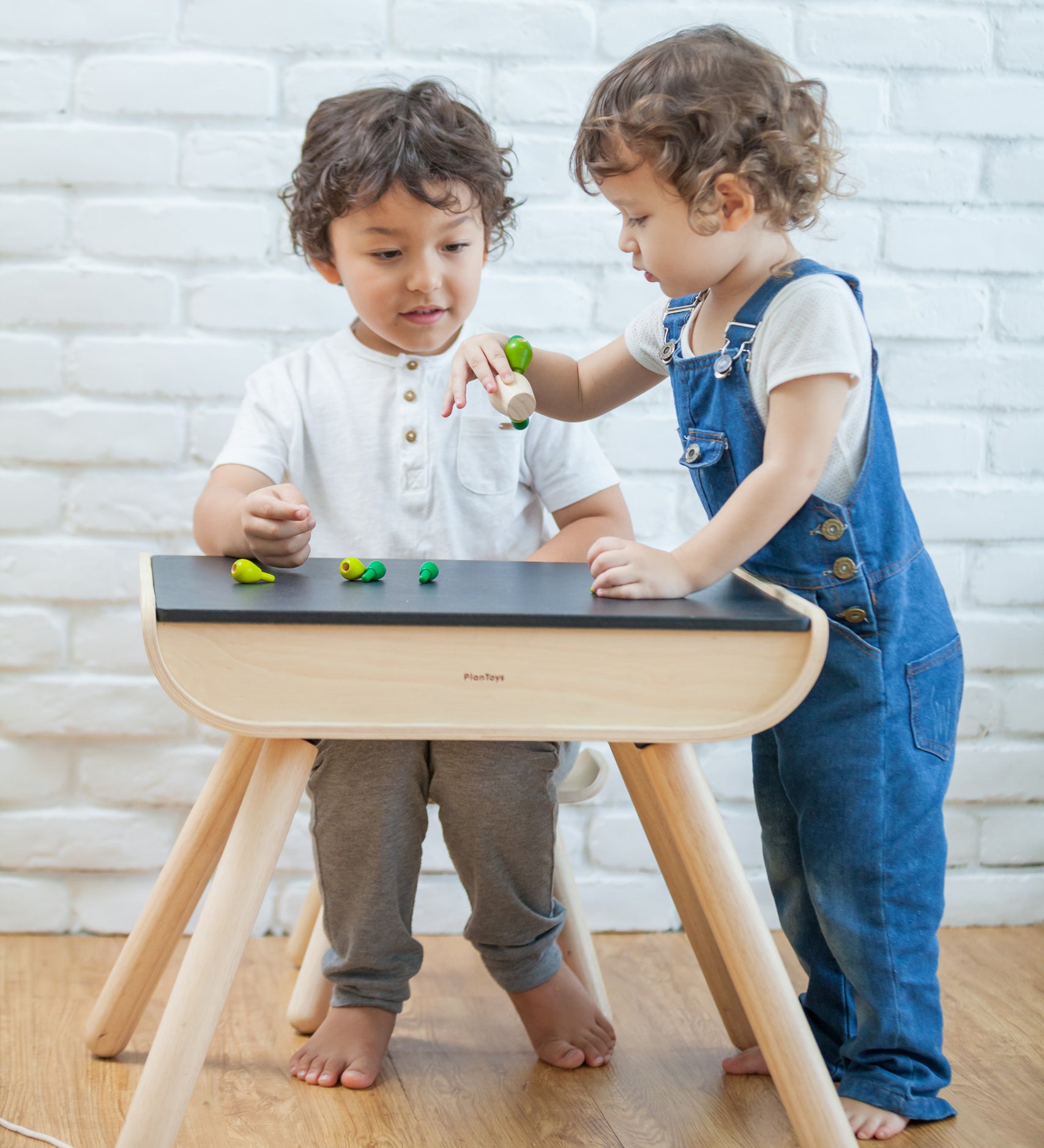 A child sitting by a PlanToys Black Table & Chair. Another child is standing by their side.