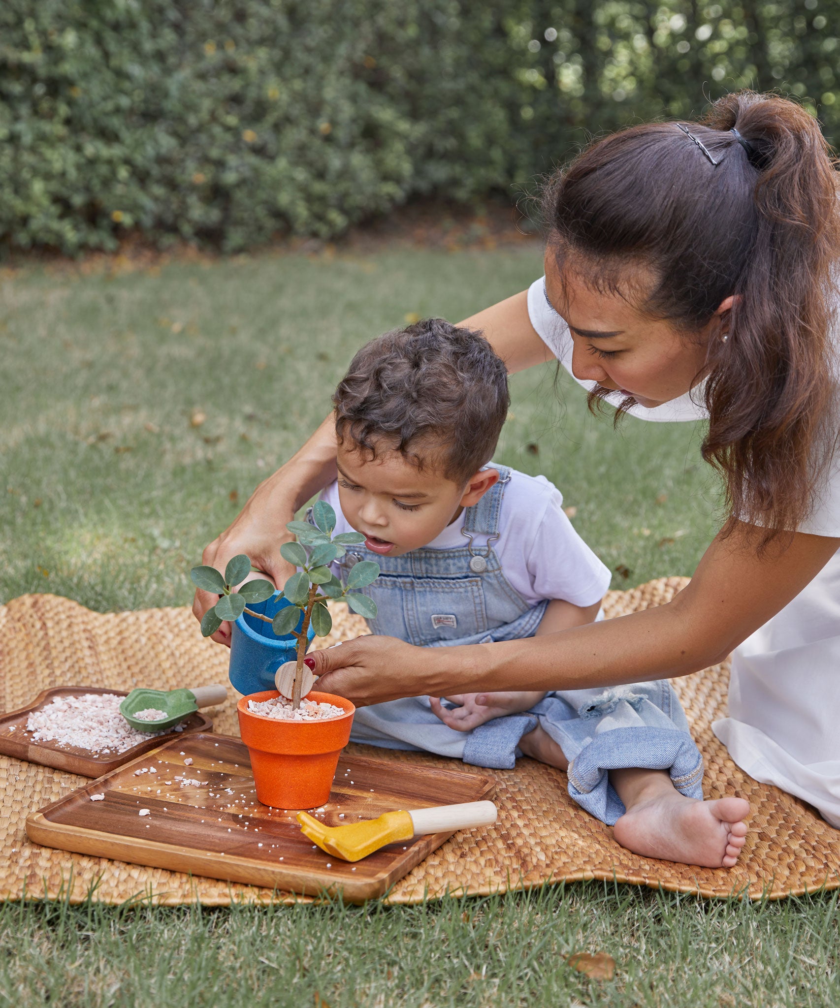 A child playing with the PlanToys Gardening Set, an adult is sitting behind them helping them use the watering can. 