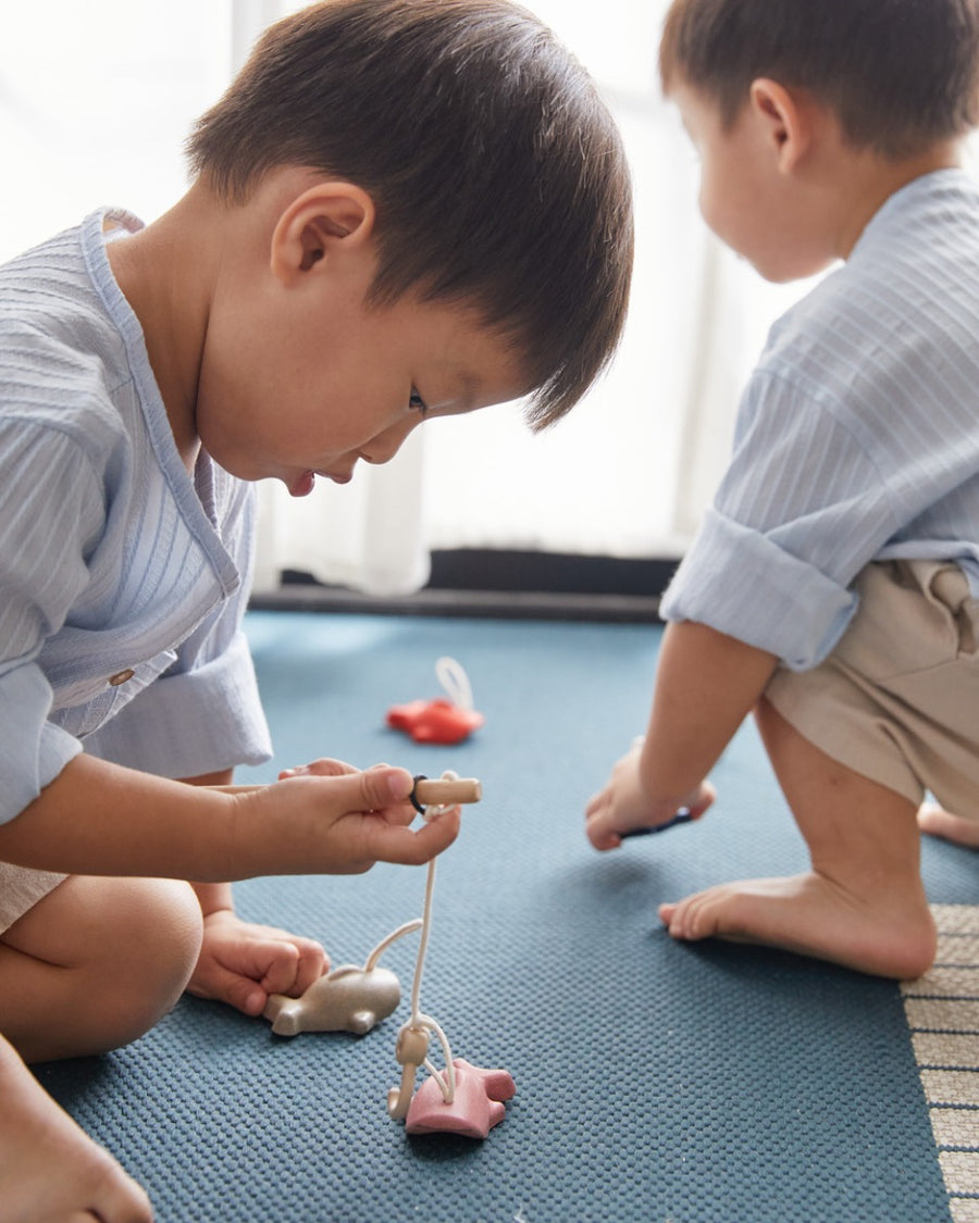 Close up of young boy crouched on a blue carpet hooking the PlanToys wooden fish toy with a toy fishing rod