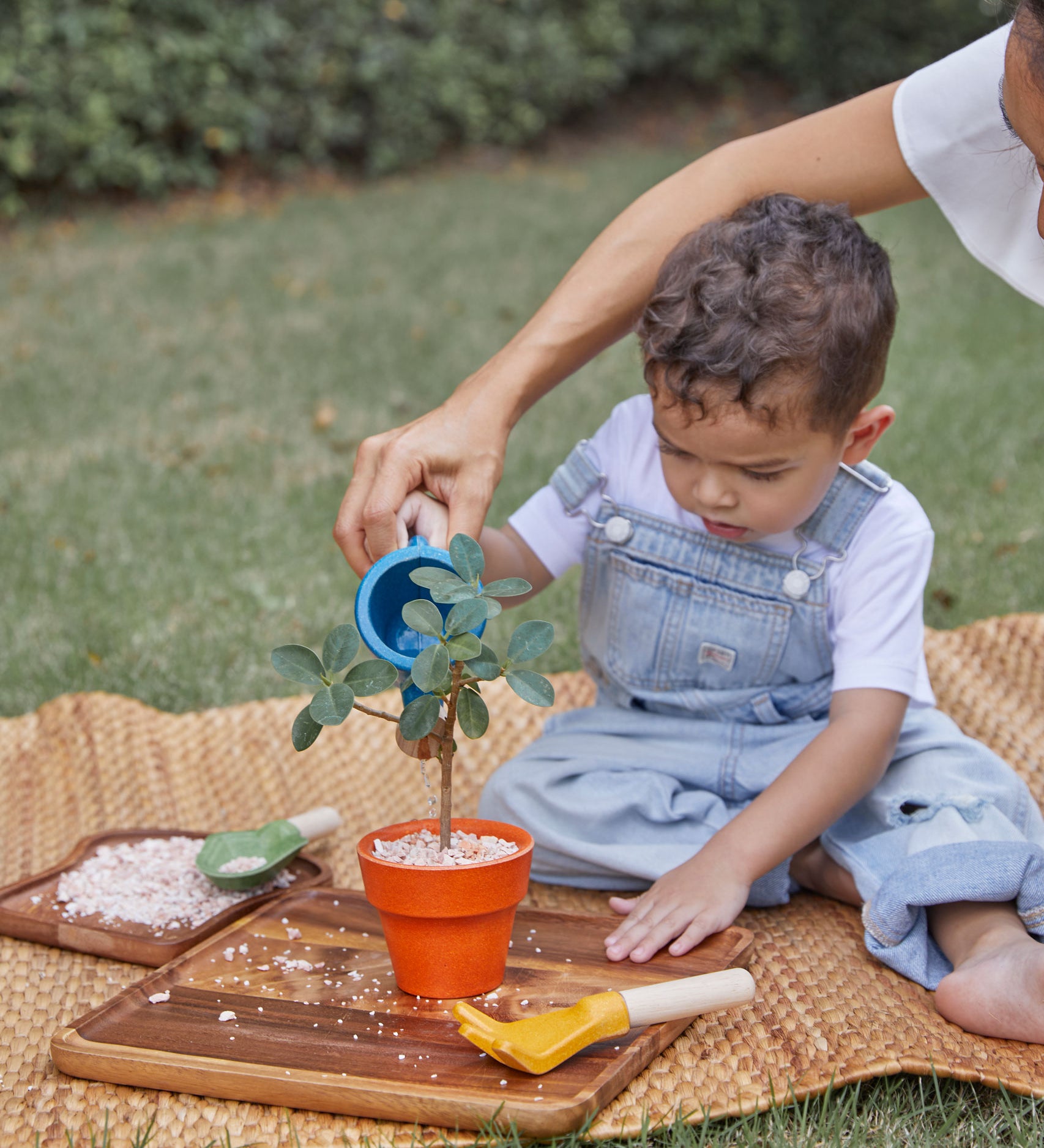 A child playing with the PlanToys Gardening Set, an adult is sitting behind them helping them use the watering can. 