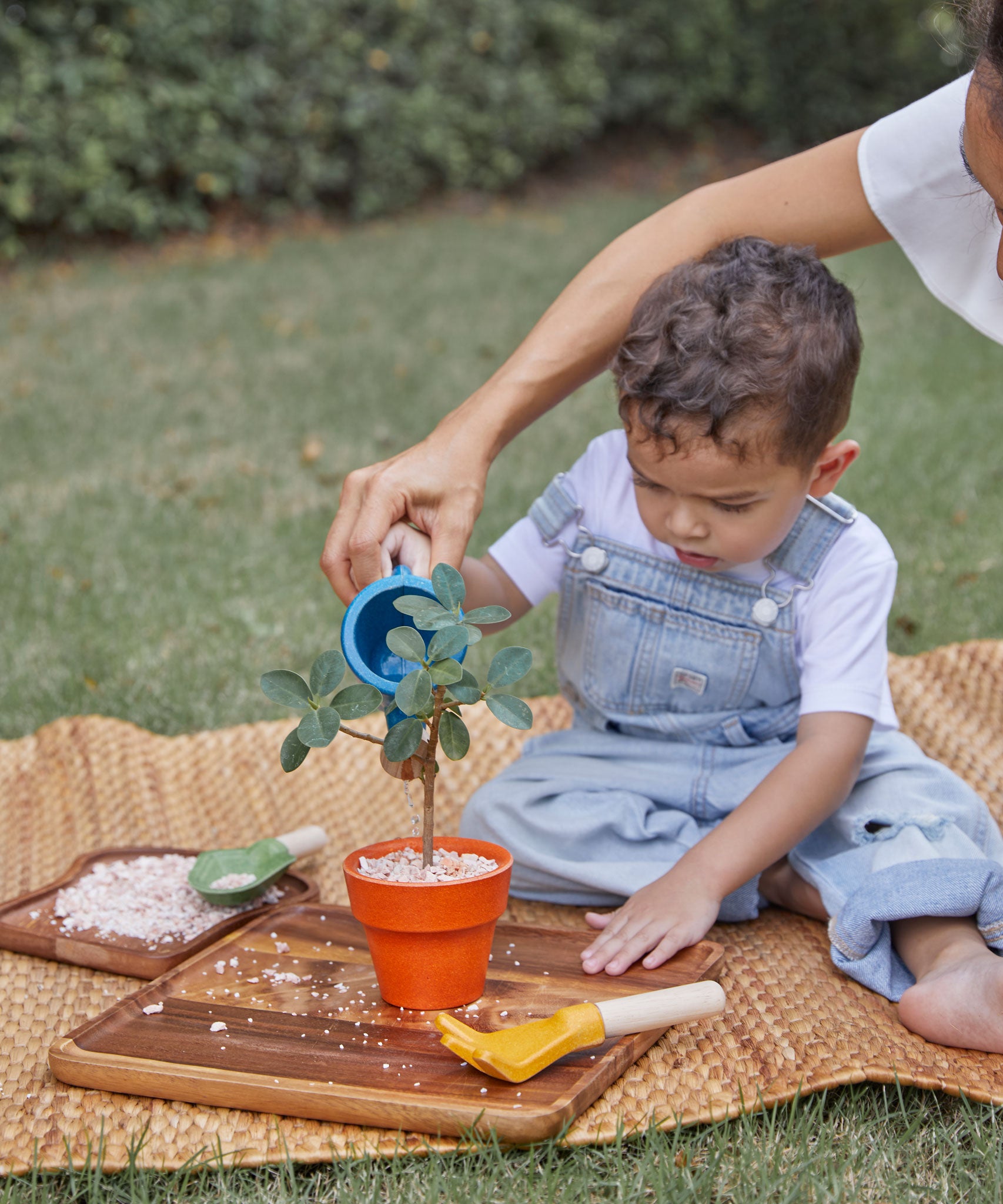 A child playing with the PlanToys Gardening Set, an adult is sitting behind them helping them use the watering can. 