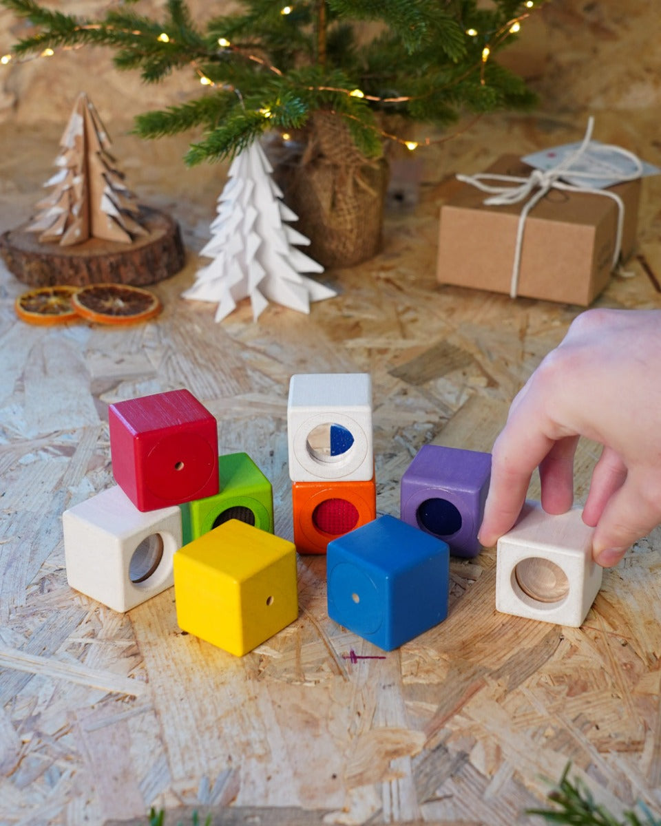 Close up of hand grabbing a white block from the PlanToys kids wooden sensory activity blocks in front of a small Christmas tree.