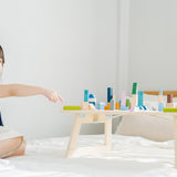 Young girl sat on a white bed pointing at the PlanToys eco-friendly wooden city blocks toy set