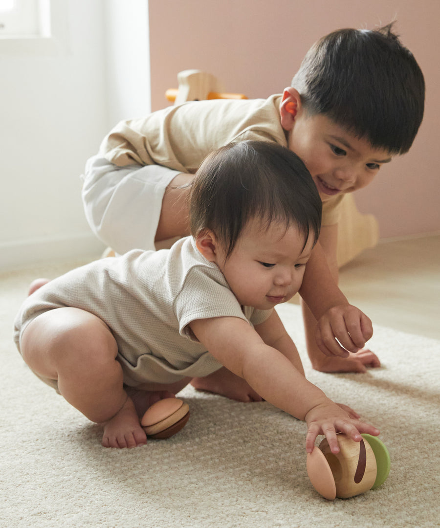 Two young children crawling on the floor reaching for the PlanToys clapping roller sensory toy. 