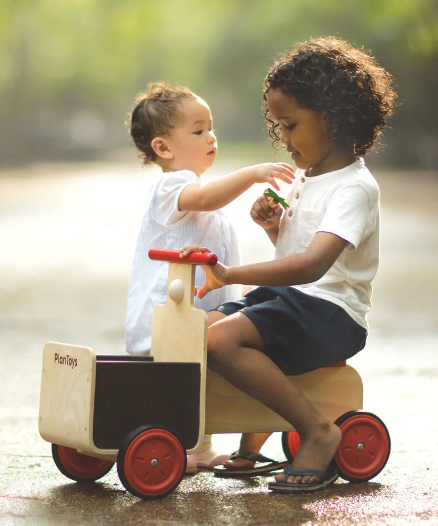 A child sitting on the PlanToys wooden delivery bike ride on toy. Another child is sstanding besides the delivery bike. 