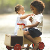 A child sitting on the PlanToys wooden delivery bike ride on toy. Another child is sstanding besides the delivery bike. 