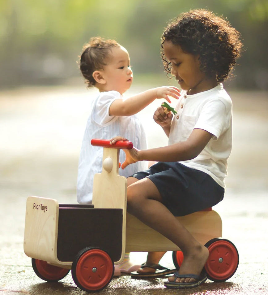 A child sitting on the PlanToys wooden delivery bike ride on toy. Another child is sstanding besides the delivery bike. 