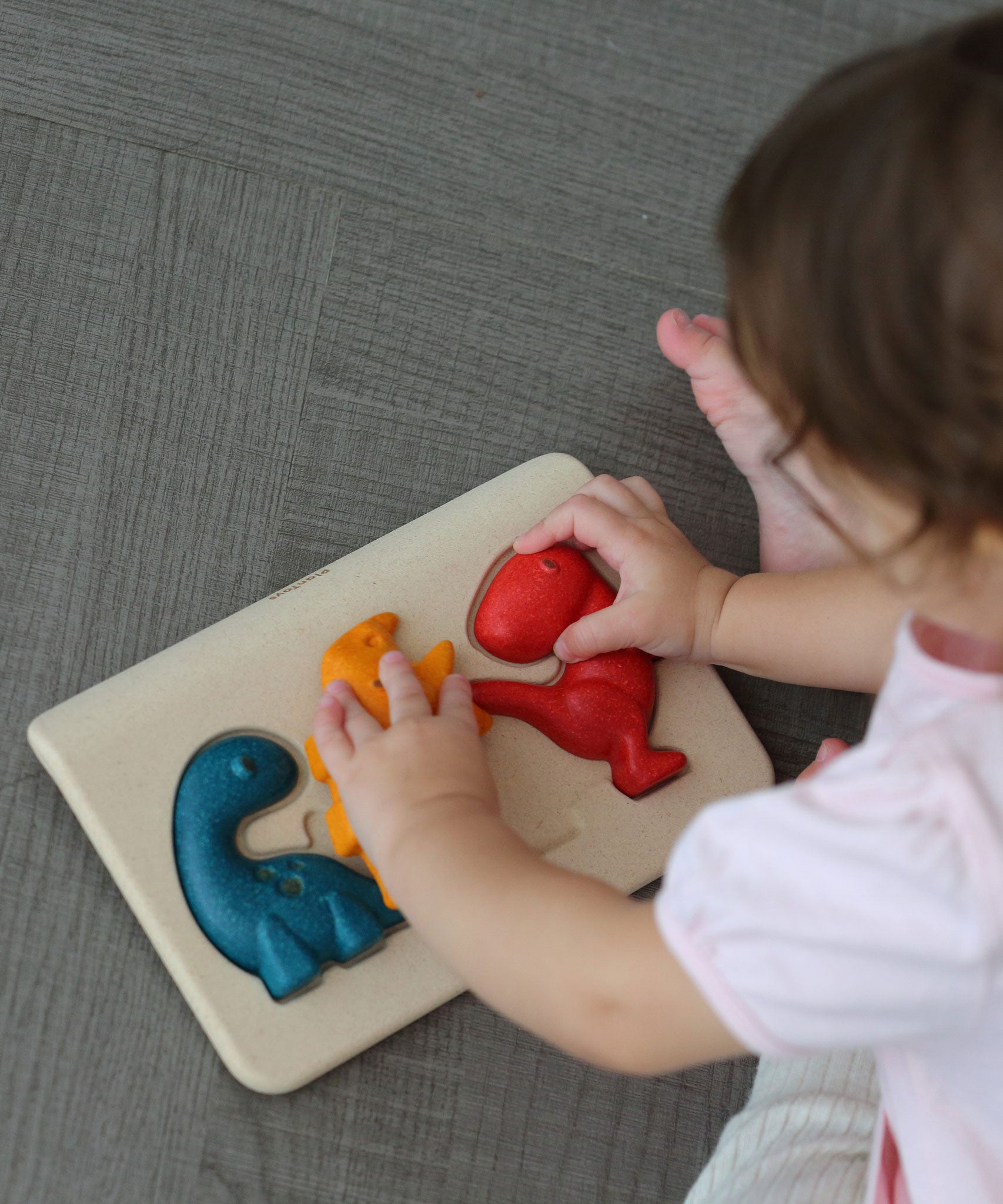 An overhead view of a child playing with the PlanToys Dino Puzzle, They have their hands on the orange and red dinosaur pieces. 