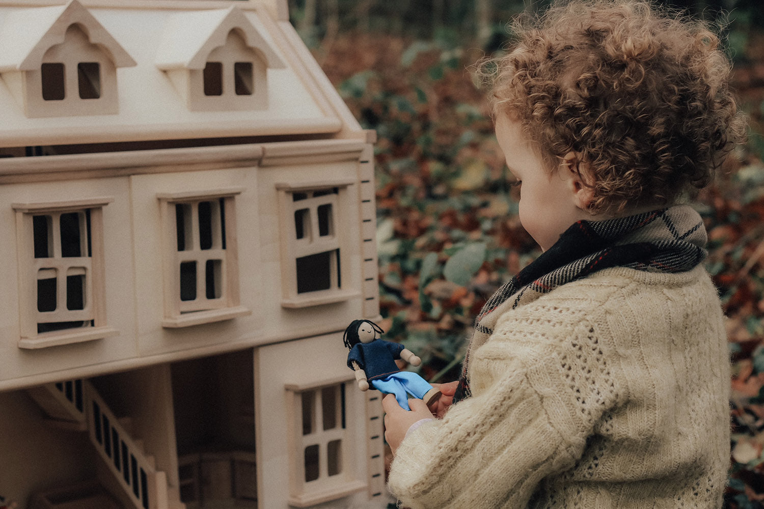 A child playing with the PlanToys Victorian Dolls House in a forest. They are holing onto a wooden dolly. 