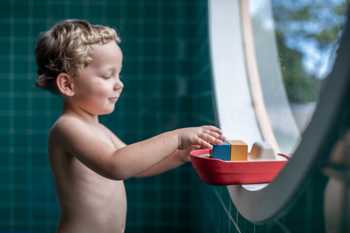 Boy stood in a round window playing with the PlanToys eco-friendly wooden cargo ship toy