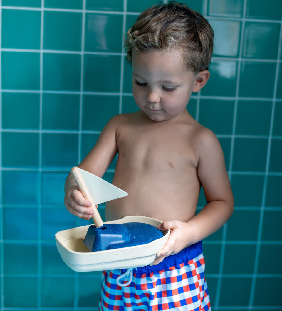 Close up of a young boy holding the PlanToys solid wooden sail boat toy in front of some blue tiles