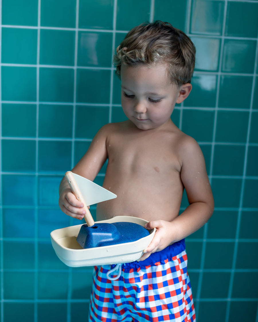 Close up of a young boy holding the PlanToys solid wooden sail boat toy in front of some blue tiles