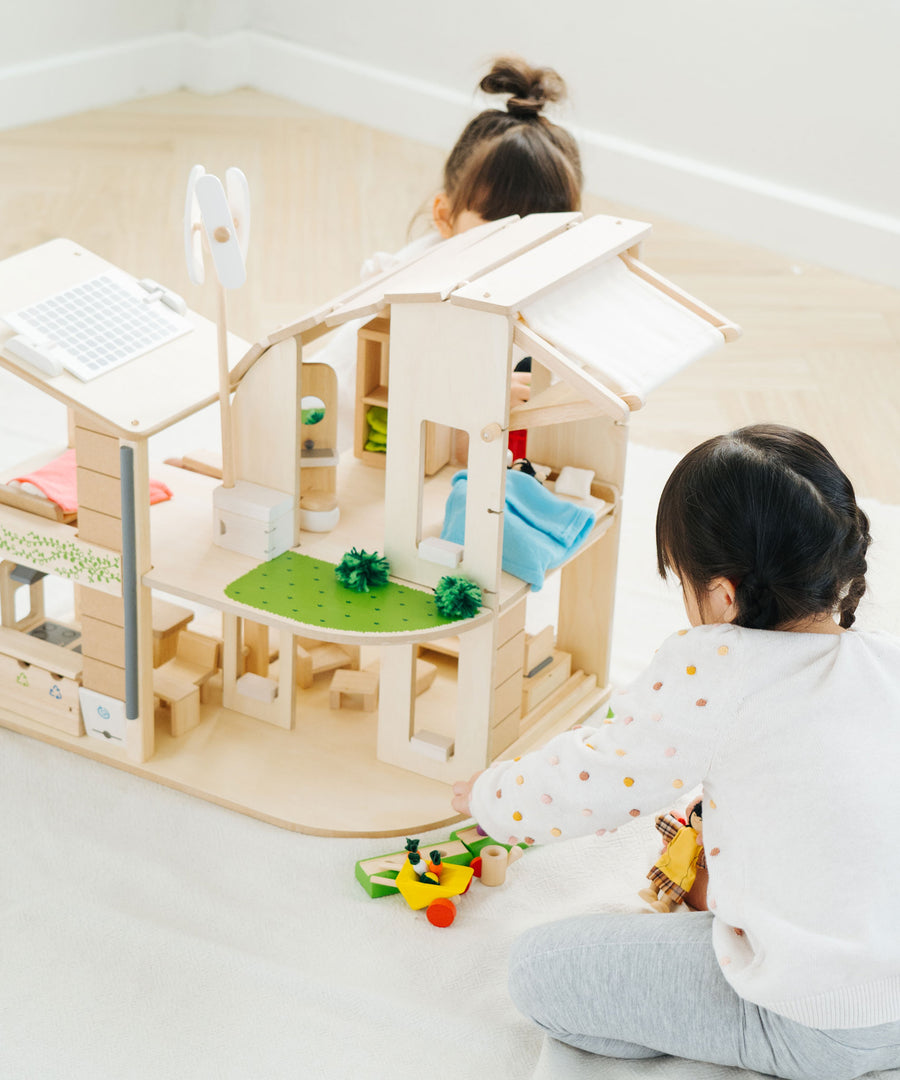 Two children playing with the PlanToys Green Dolls' House and Furniture Set. The house is placed on a white coloured rug and the children are sitting on opposite sides of the house. 