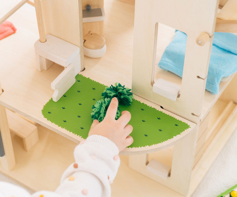 A close up of the grass effect painted areas on the side of the PlanToys Green Dolls' House and Furniture Set. A child's hand can be seen reaching for a green grass like pom pom. 