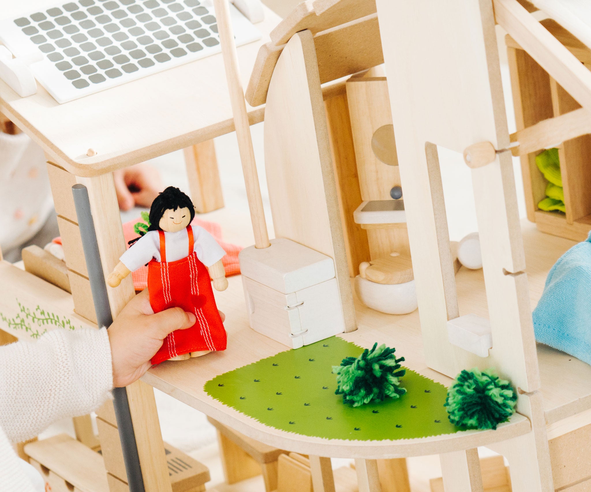 A close up of the grass effect painted areas on the side of the PlanToys Green Dolls' House and Furniture Set. A child's hand can be seen besides the grassy area holding a doll wearing a red outfit. 