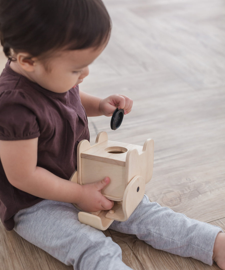 A child sitting on a wooden surface holding the PlanToys Elephant Money Bank upside down. 