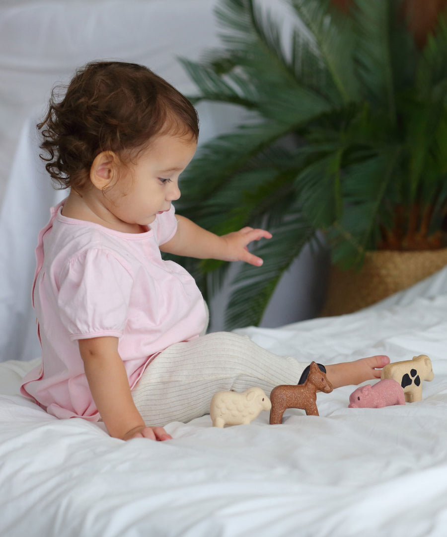 A child sitting on a white duvet playing with the figures from the  PlanToys Farm Animals Set. 
