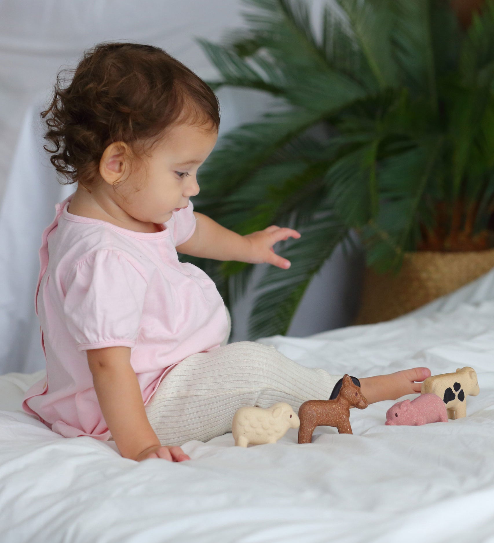 A child sitting on a white duvet playing with the figures from the  PlanToys Farm Animals Set. 
