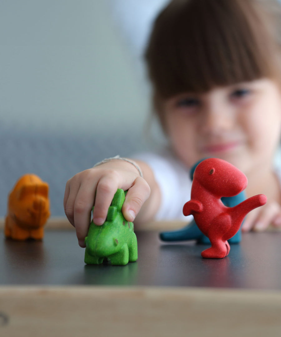 The PlanToys Dino Set on a PlanToys table, a child's hand is on the green dinosaur and the child can be seen in the background.