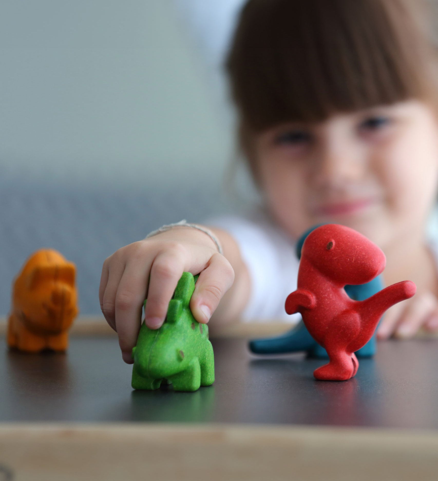 The PlanToys Dino Set on a PlanToys table, a child's hand is on the green dinosaur and the child can be seen in the background.