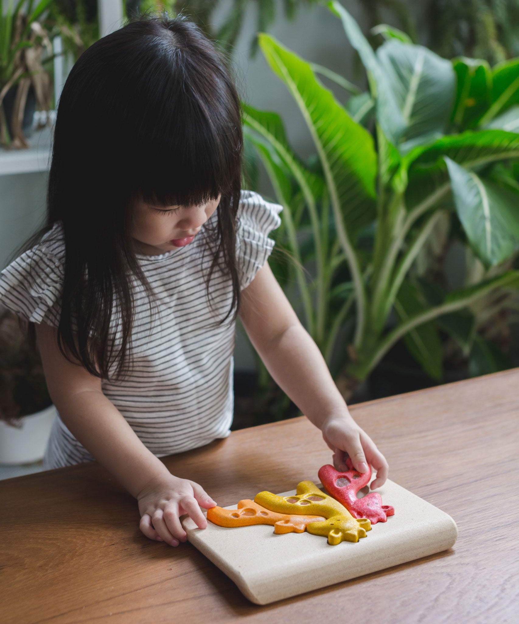 A child stood by the side of a wooden table playing with the PlanToys Giraffe Puzzle. 
