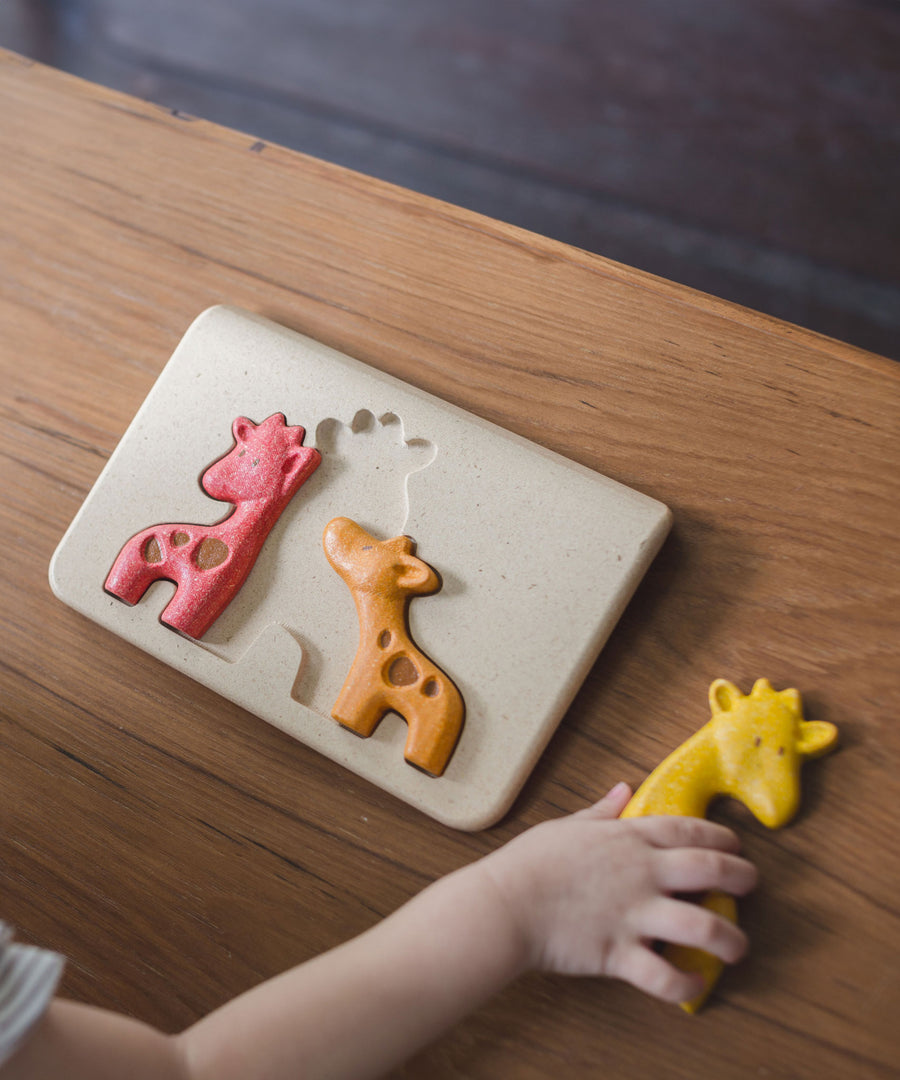 A top view of the PlanToys Giraffe Puzzle on a wooden table. A child is holding the yellow puzzle piece.