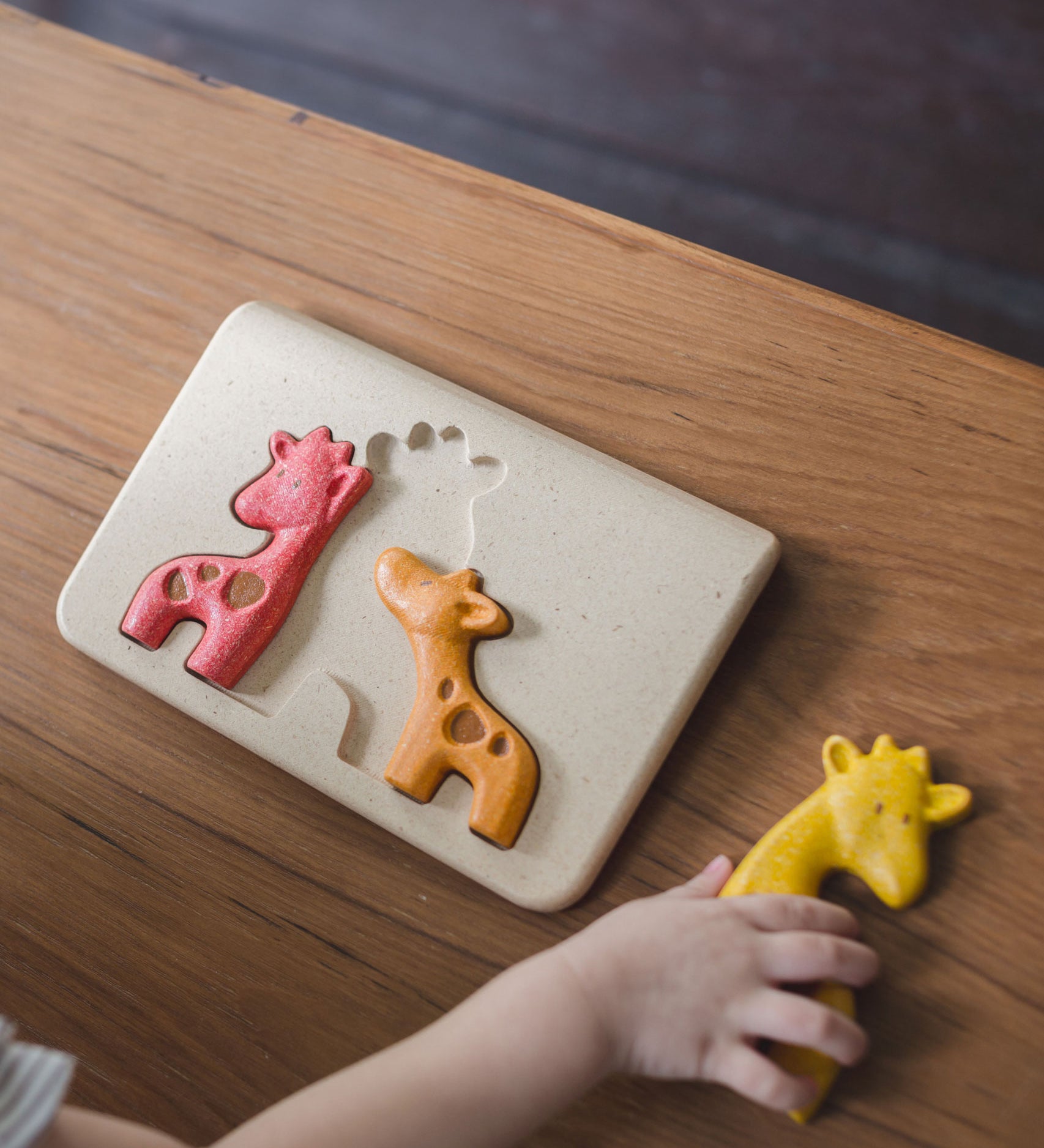 A top view of the PlanToys Giraffe Puzzle on a wooden table. A child is holding the yellow puzzle piece.