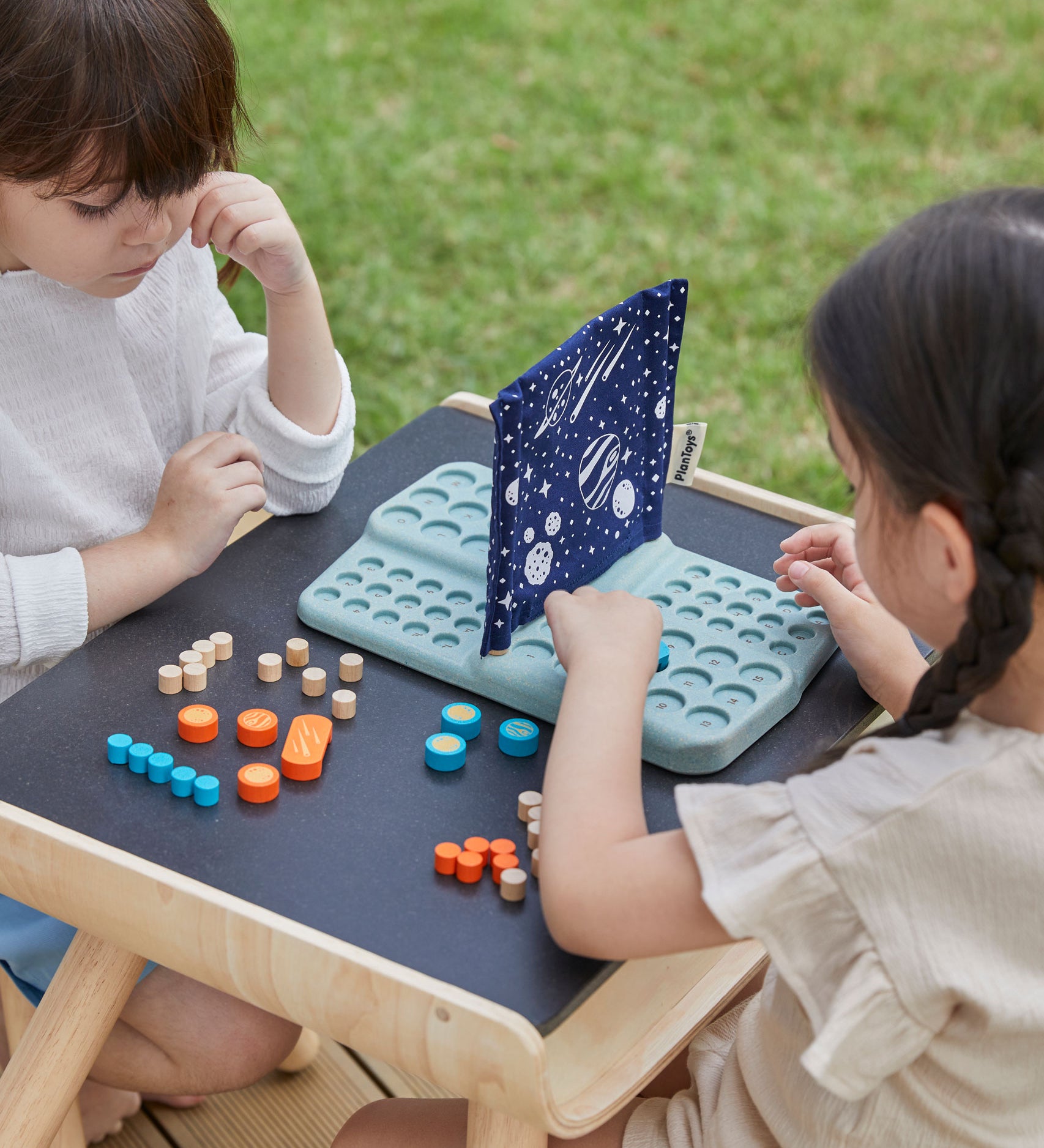 Two children sitting at opposite side of the PlanToys desk playing with the PlanToys Guess My Planet Game.
