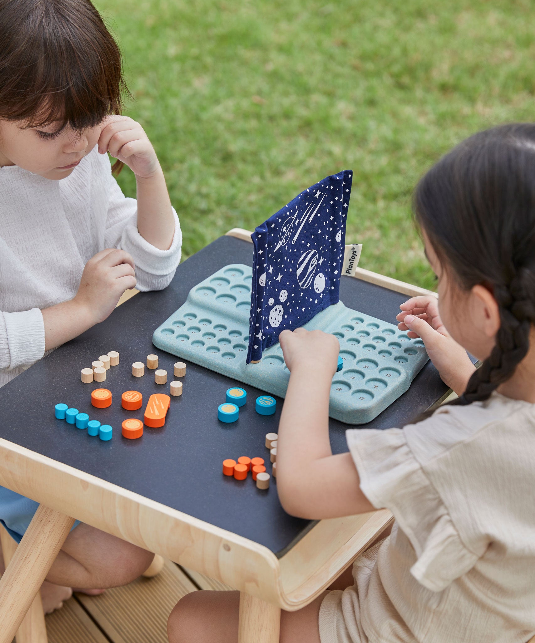 Two children sitting at opposite side of the PlanToys desk playing with the PlanToys Guess My Planet Game.
