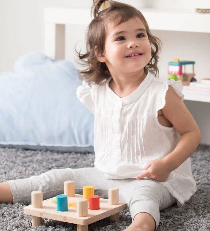 A young child sitting on a grey coloured rug playing with the PlanToys Hammer Peg wooden toy. 