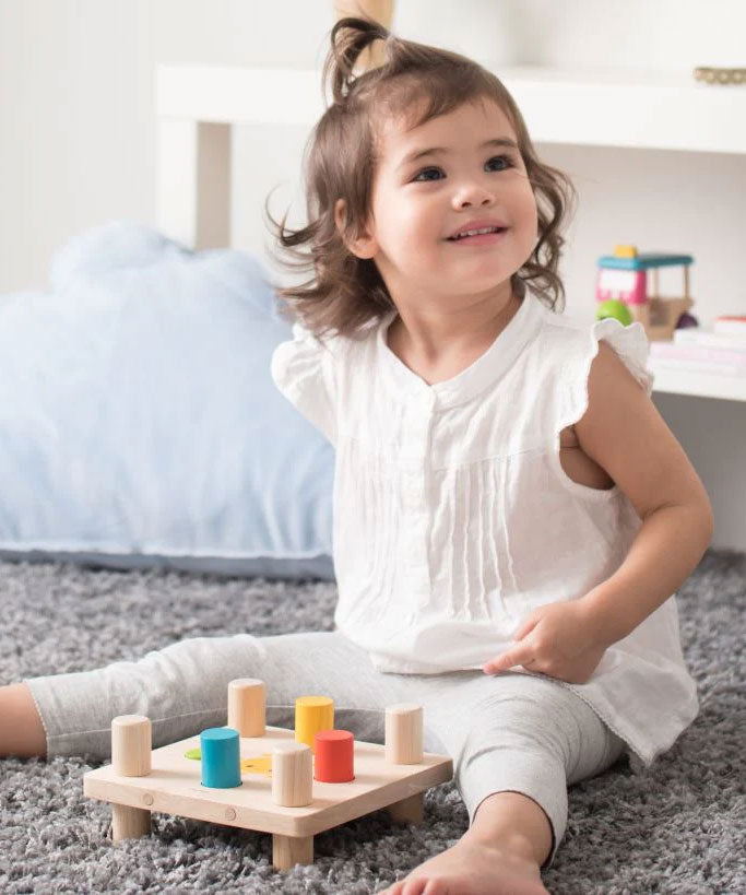 A young child sitting on a grey coloured rug playing with the PlanToys Hammer Peg wooden toy. 