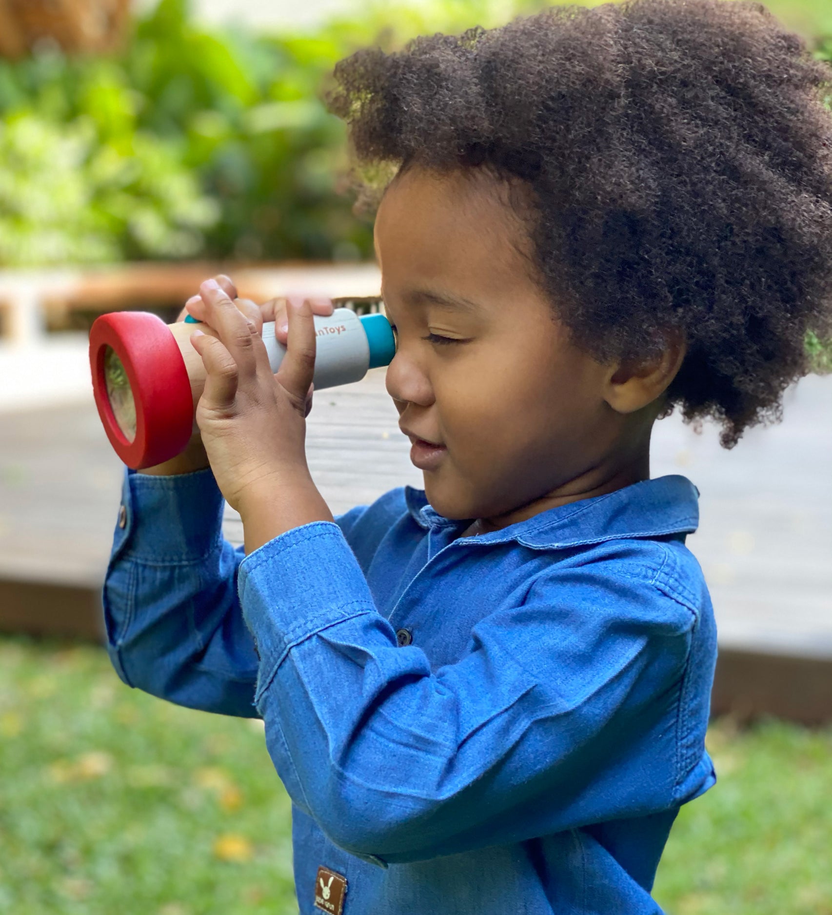 A child playing with the PlanToys Kaleidoscope toy. The child is holding up the PlanToys Kaleidoscope to their eye.  