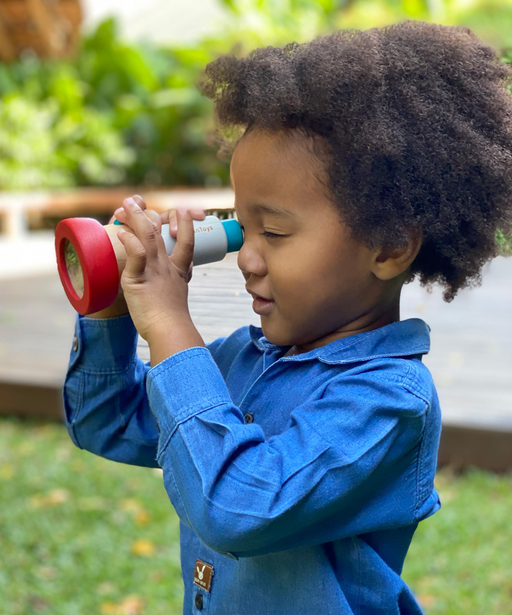 A child playing with the PlanToys Kaleidoscope toy. The child is holding up the PlanToys Kaleidoscope to their eye.  