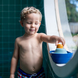 Young boy stood in a round window playing with the PlanToys eco-friendly wooden tugboat