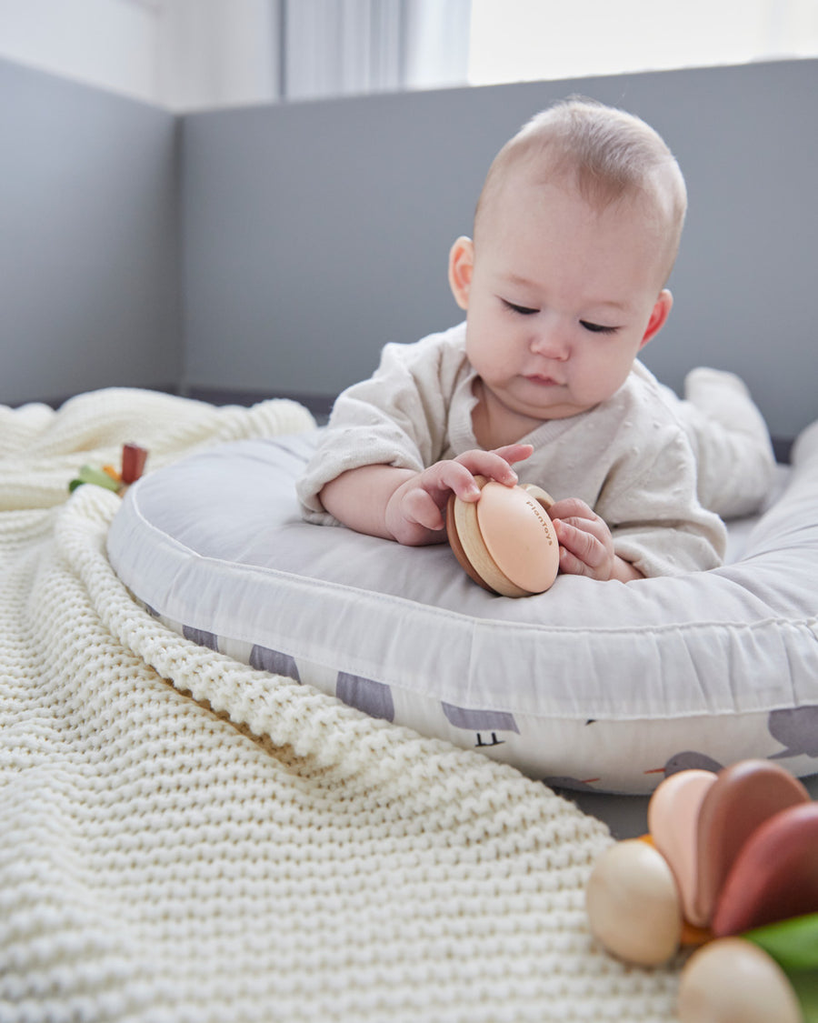 Baby laying down on a white cushion playing with the PlanToys wooden Shake N Rattle toy