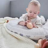 Baby laying down on a white cushion playing with the PlanToys wooden Shake N Rattle toy