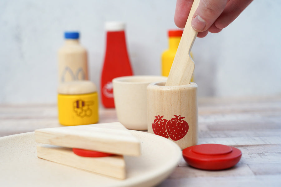 Close up of a hand holding a PlanToys kids wooden knife in a wooden pot of PlanToys strawberry jam play food on a grey background