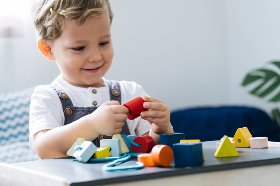 Close up of a child sat down threading PlanToys wooden lacing blocks onto a string