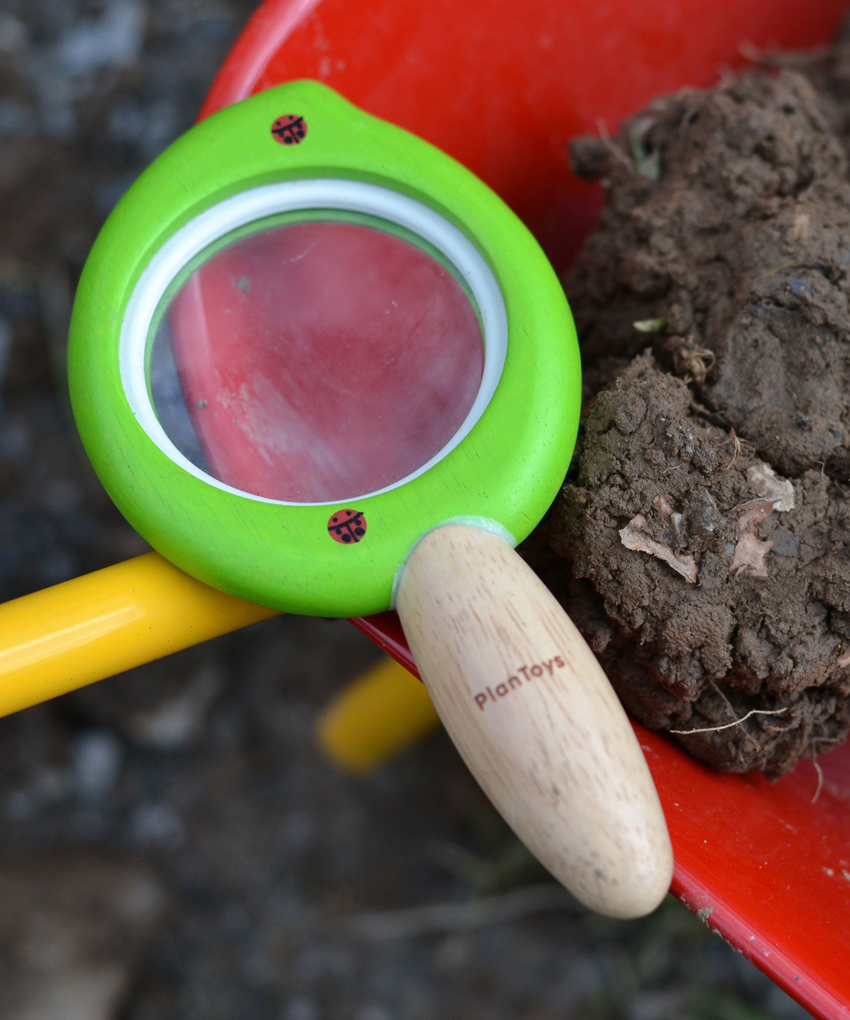 A close up of a Plan Toys Leaf Magnifier toy placed on a side of a red wheelbarrow which is filled with mud. 