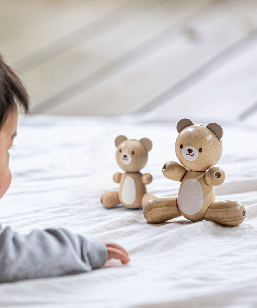 A PlanToys Bear next to the smaller bear on a light coloured material, a child can be seen in the corner looking towards the bears.