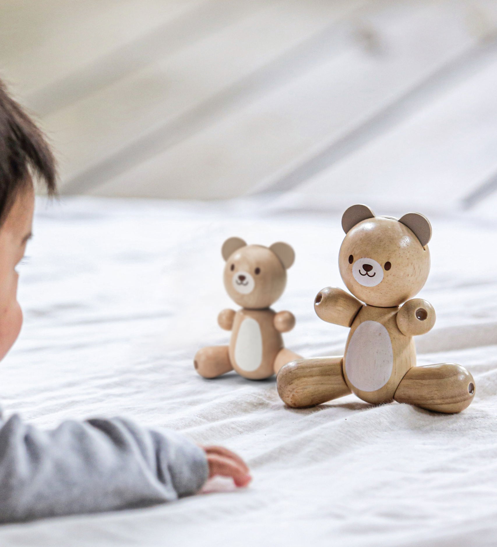 A PlanToys Bear next to the smaller bear on a light coloured material, a child can be seen in the corner looking towards the bears.