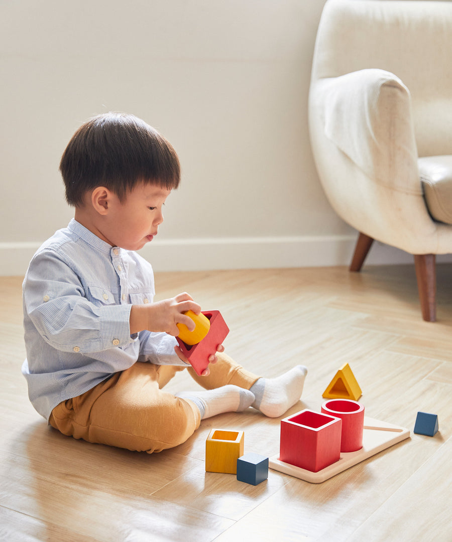 A child sitting on a wooden floor playing with the PlanToys Matching & Nesting Shapes Puzzle.